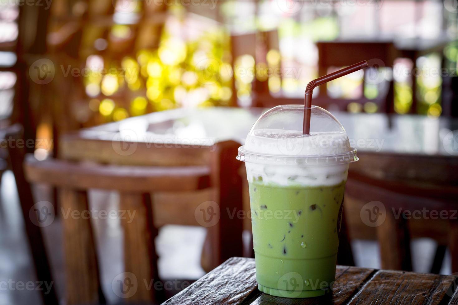 Iced green tea or matcha latte with milk foam in a plastic cup with brown straw on a wooden table in the coffee shop with sun light. Healthy drink and beverage concept. photo