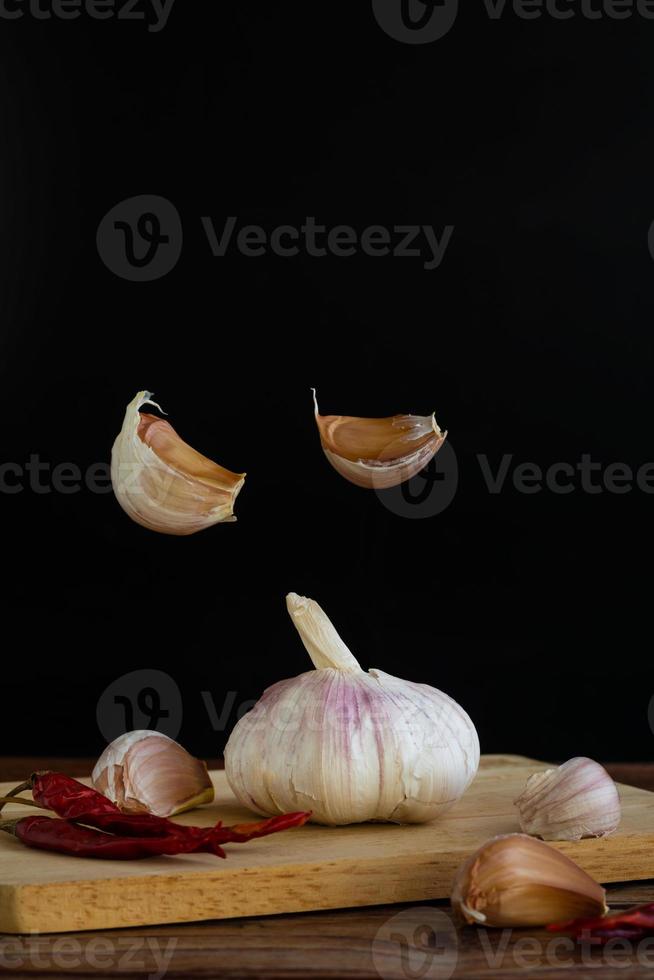 grupo de ajo en la tabla de cortar y algunos dientes de ajo flotando en el aire y ají seco rojo sobre una mesa de madera con fondo negro. copie el espacio para su texto. foto