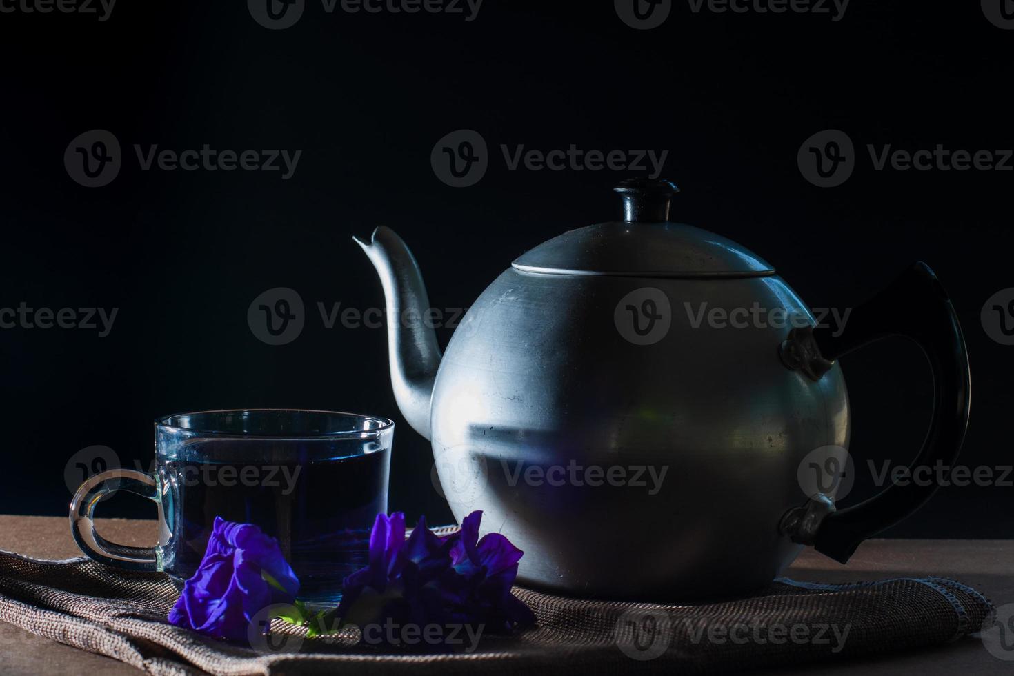 Set of Cup and metal pot of Butterfly pea tea with fresh violet flower on brown tablecloth and wooden table on black background. Healthy beverage for drink. Herbs and medical concept. photo