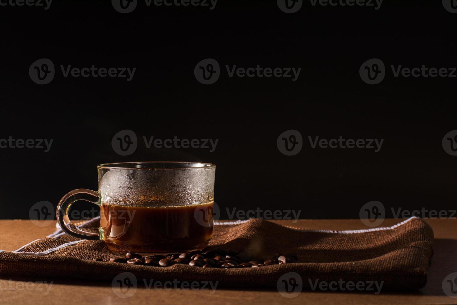 Cup of hot coffee with smoke and group of coffee beans on brown table cloth with black background with copy space for your text. Benefits of coffee concept. photo