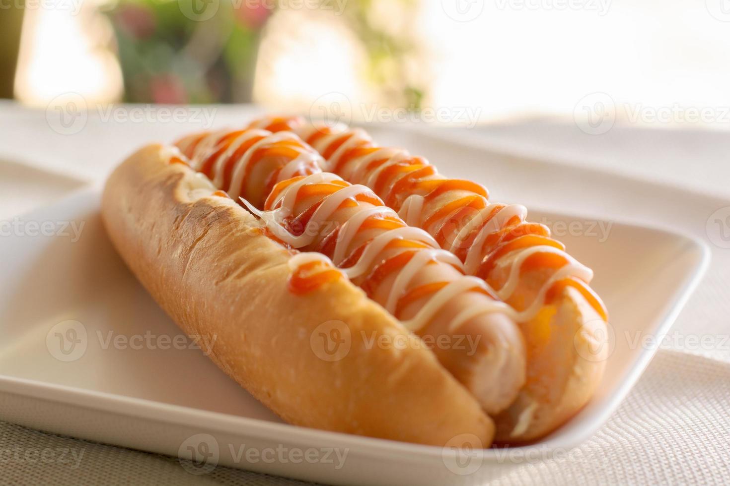 Hot dog with bun and ketchup, mayonnaise on white plate. Sausage sandwich for lunch. Fast food concept. photo