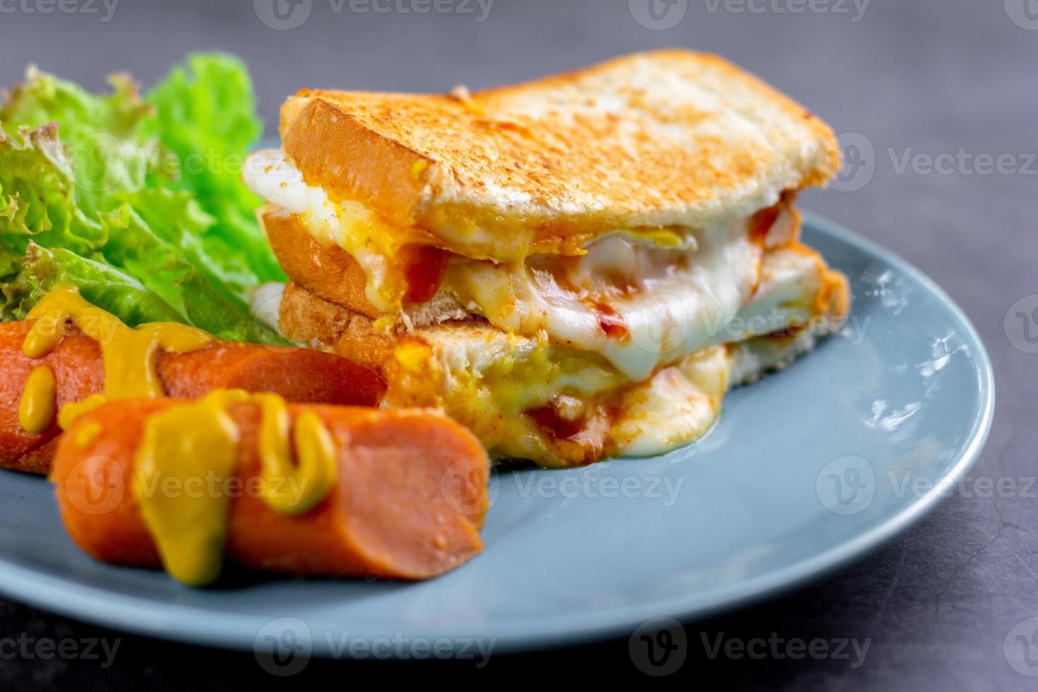American breakfast concept. Sandwich with melting cheese and blurry sausage and fresh lettuce on a blue plate on a dark rock table. Homemade food in the morning. photo