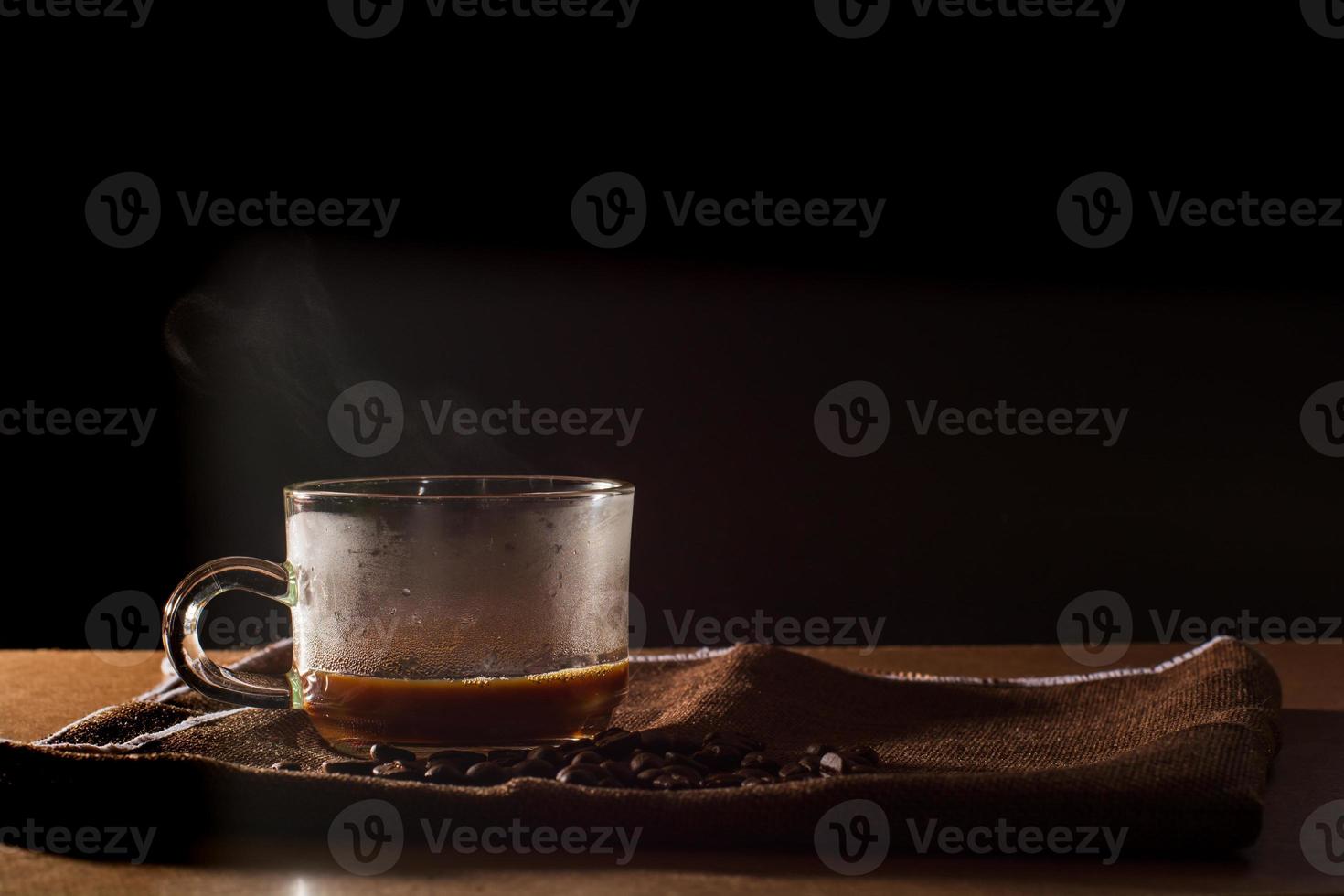 Cup of hot coffee with smoke and group of coffee beans on brown table cloth with black background with copy space for your text. Benefits of coffee concept. photo