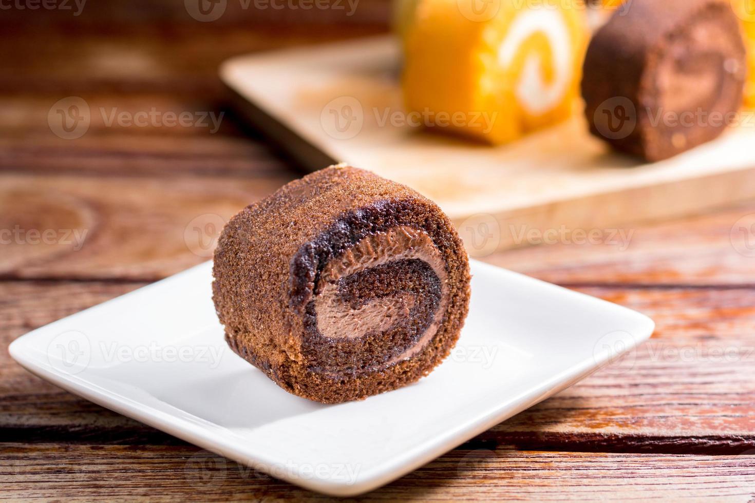 Close up delicious chocolate roll cake with whipping cream in a square white plate on a wooden table at the home kitchen with a black background and copy space. Homemade bakery concept. photo