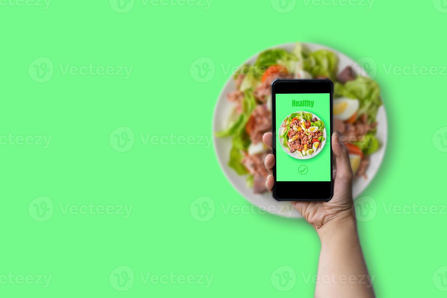 Healthy food concept. Hand holding smartphone with green text healthy with tuna salad, boil egg, lettuce and tomato on white plate on green background with copy space. photo