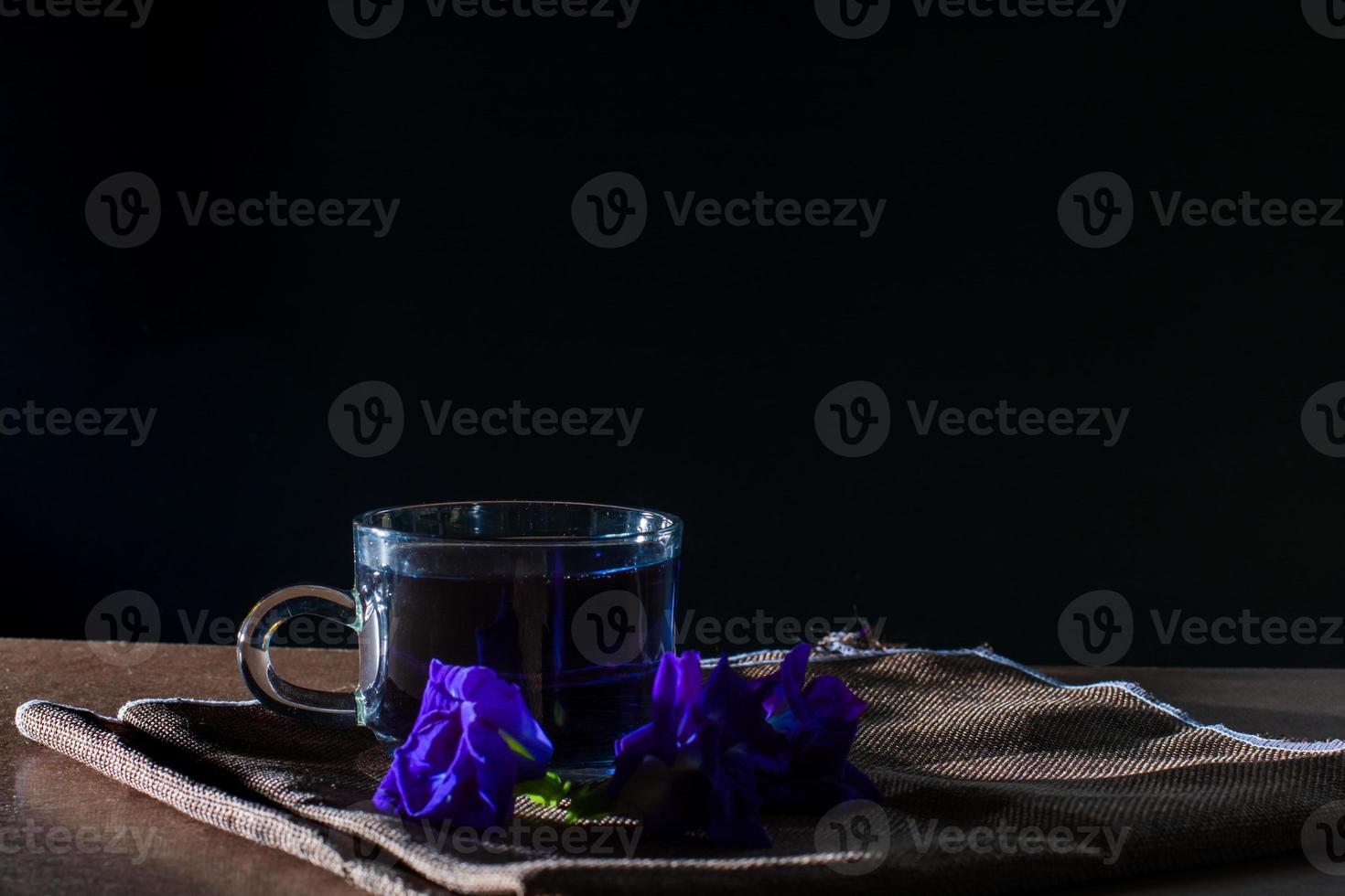 Cup of Butterfly pea tea with fresh violet flower on brown tablecloth and wooden table on black background. Healthy beverage for drink. Herbs and medical concept. photo