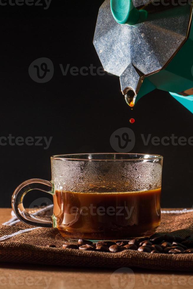 Pour hot of black coffee from green moka pot to clear coffee cup with smoke and coffee beans on brown table cloth and wooden table. Benefit of coffee concept. photo