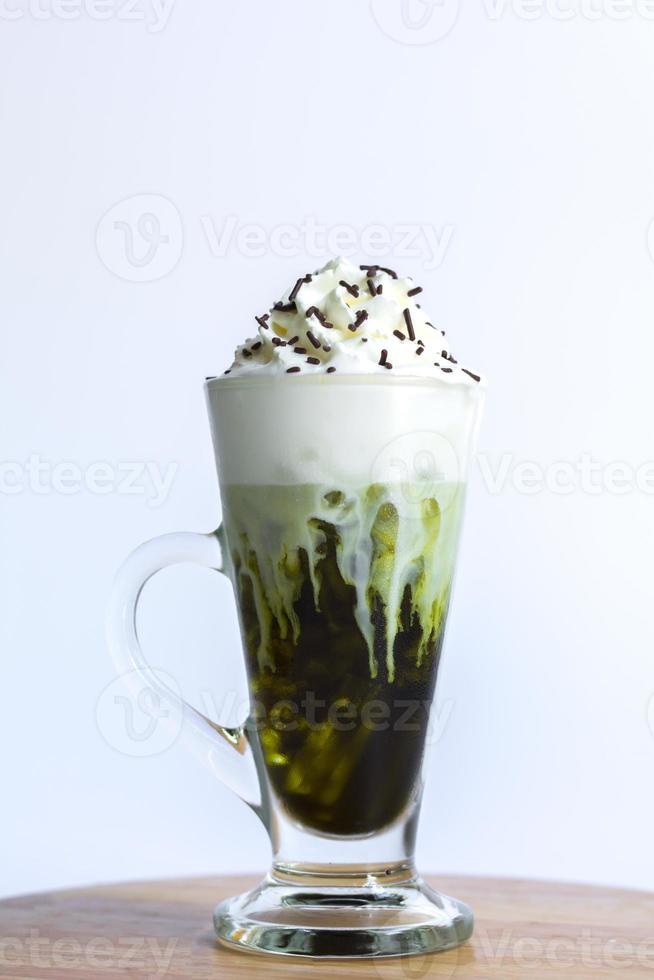 Iced green tea or matcha latte with milk foam and whipping-cream in a tall glass on a wooden table and white background. Healthy drink and beverage concept. photo