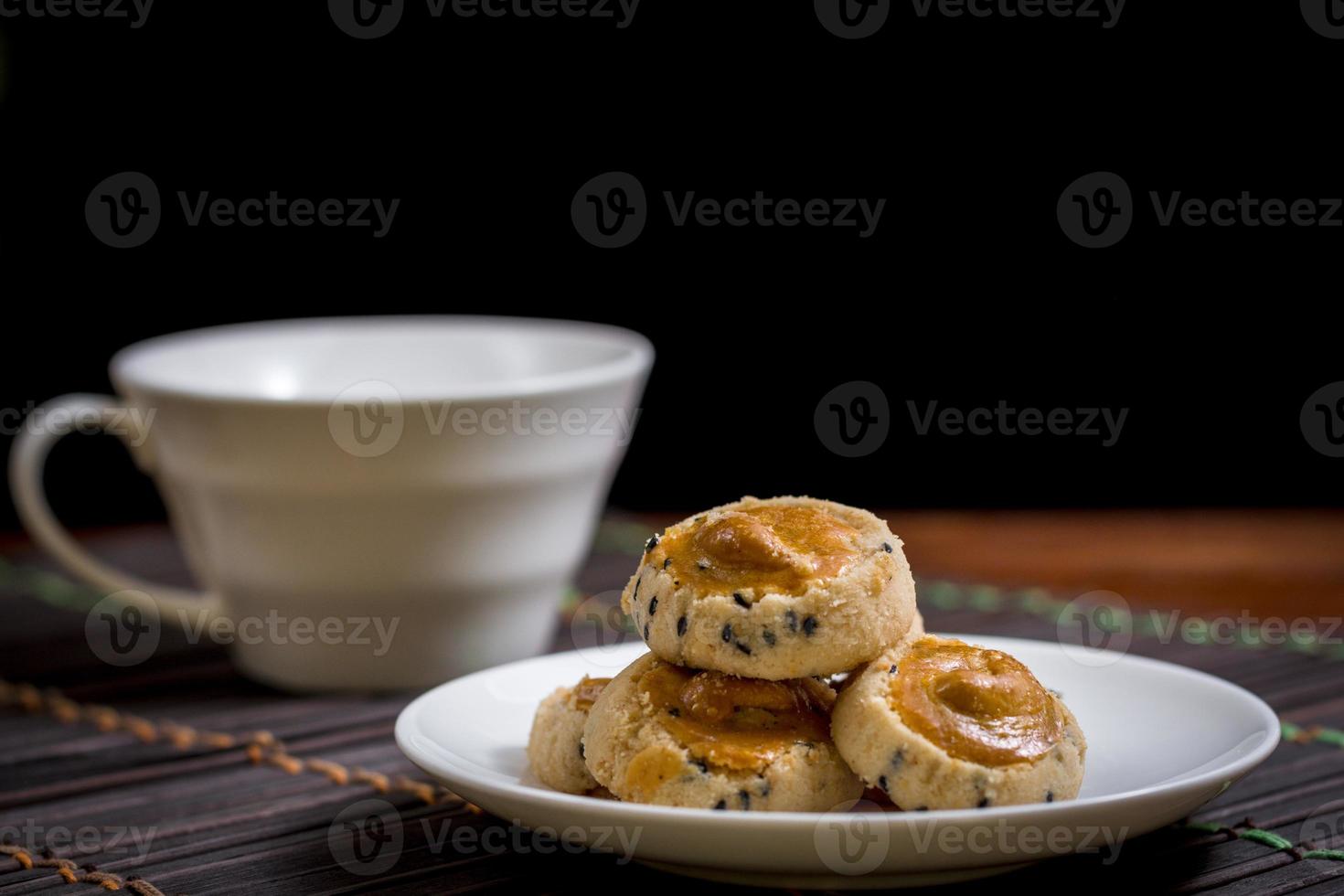 Cashew nut and black seasame cookies on white dish and wooden table with blurry cup of coffee on black background. Coffee time concept. photo