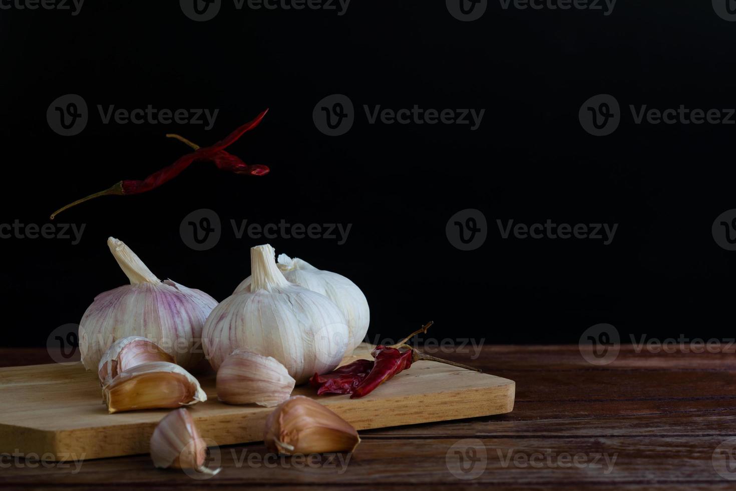 Group of garlic on chopping board and some garlic cloves floating in the air and red dried chilli on wooden table with black background. Copy space for your text. photo