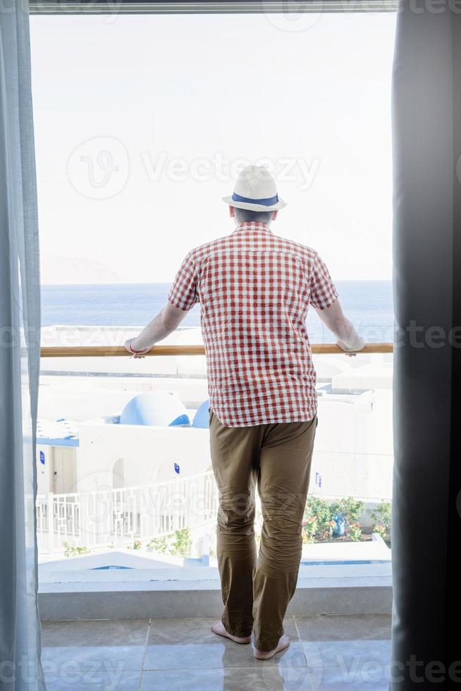 apuesto joven caucásico parado en el balcón del hotel, mirando la vista al mar foto