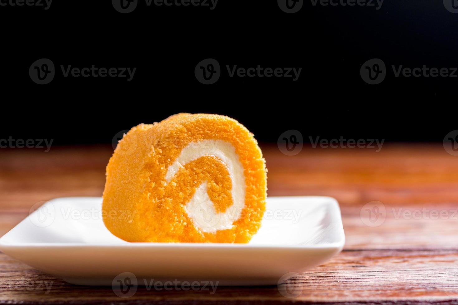 Close up delicious orange roll cake with whipping cream in a square white plate on a wooden table at the home kitchen with a black background and copy space. Homemade bakery concept. photo