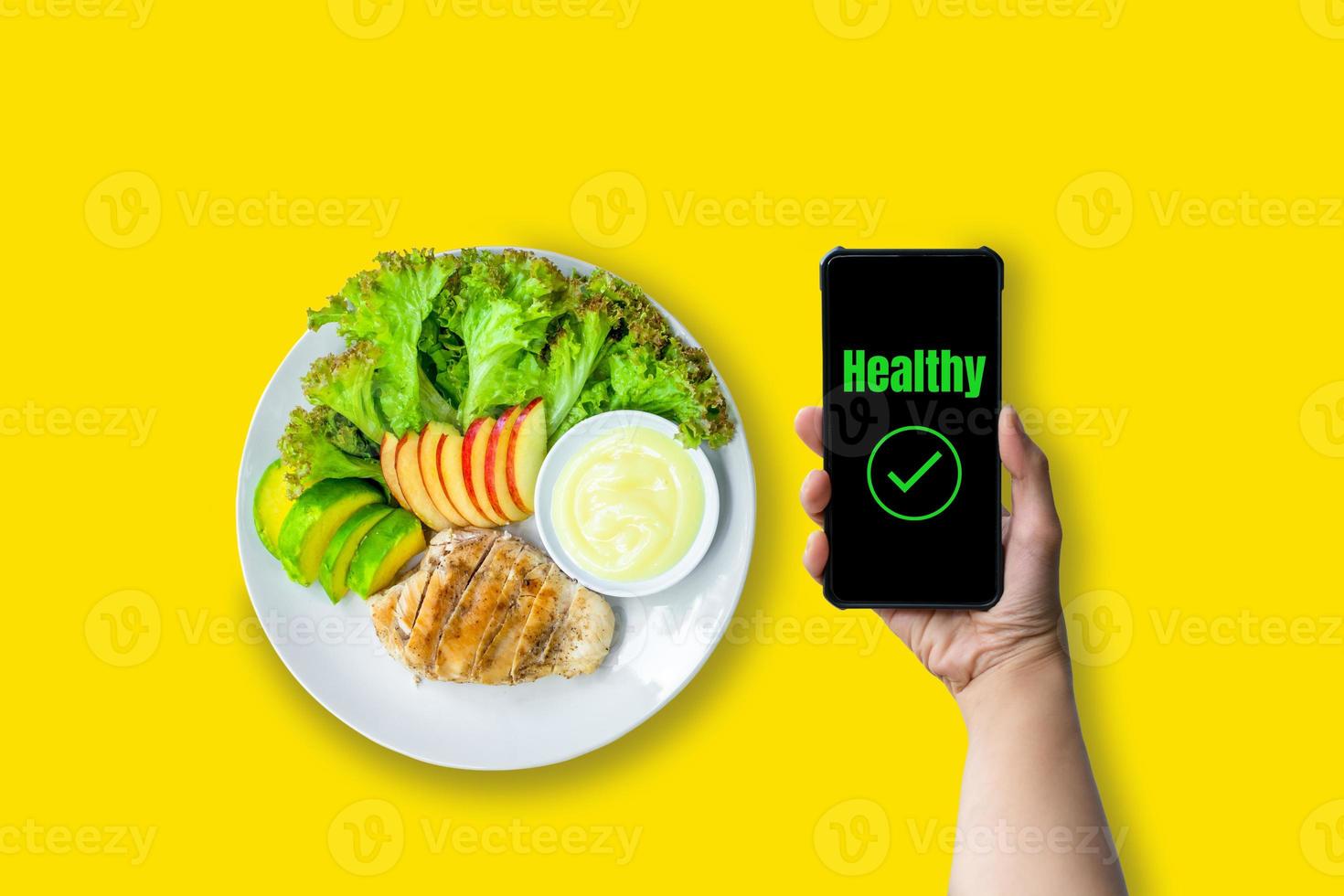 Healthy food concept. Hand holding smartphone with green text healthy with chicken salad and fresh lettuce, avocado, and apple sliced on white plate served with salad dressing on yellow background. photo
