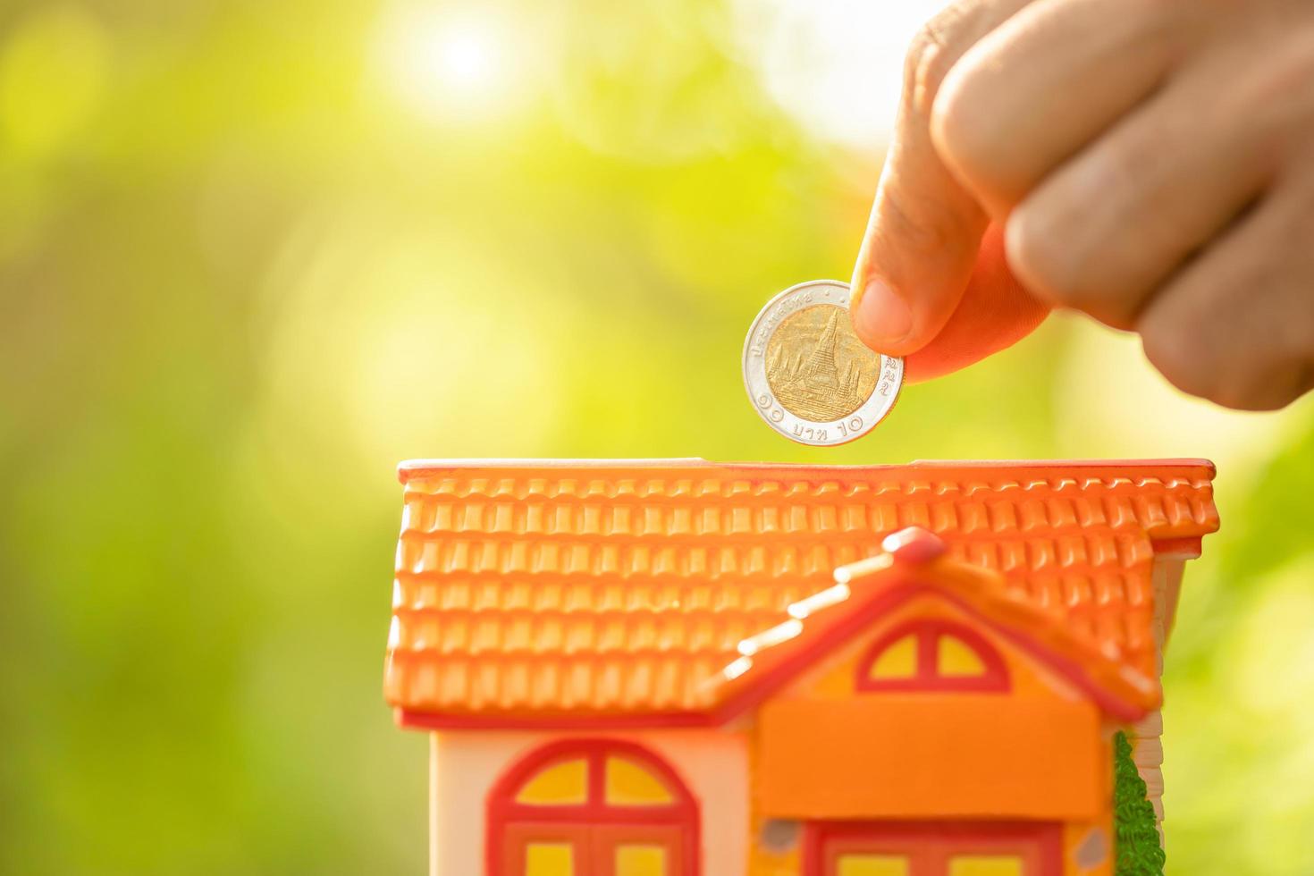 Hand put the coin to piggy bank Home Model on green nature blur background. Money savings concept photo