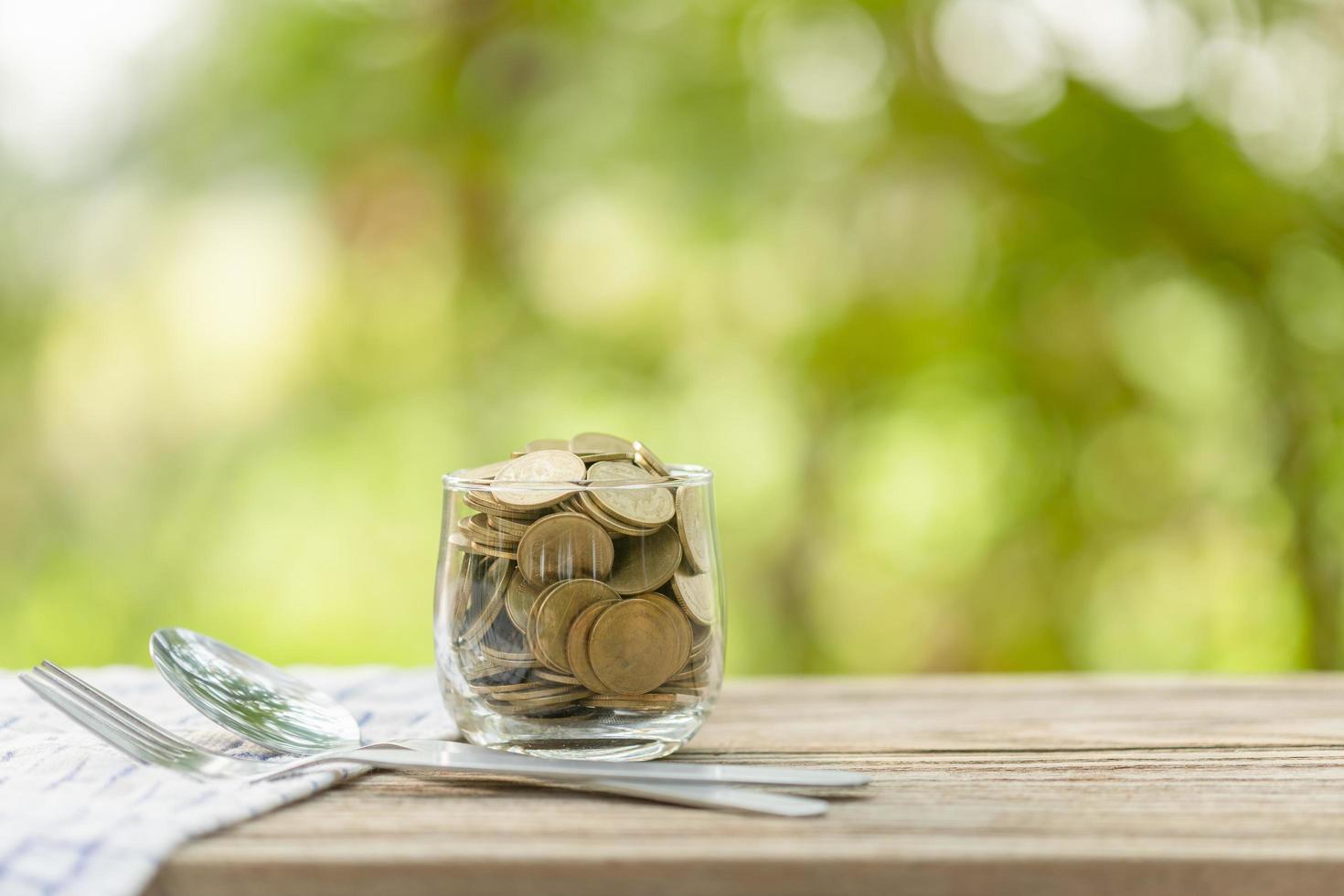 monedas en un tarro de dinero transparente, tenedor y cuchara sobre una mesa de madera con fondo de luz verde borroso. ahorro de dinero para comer concepto foto