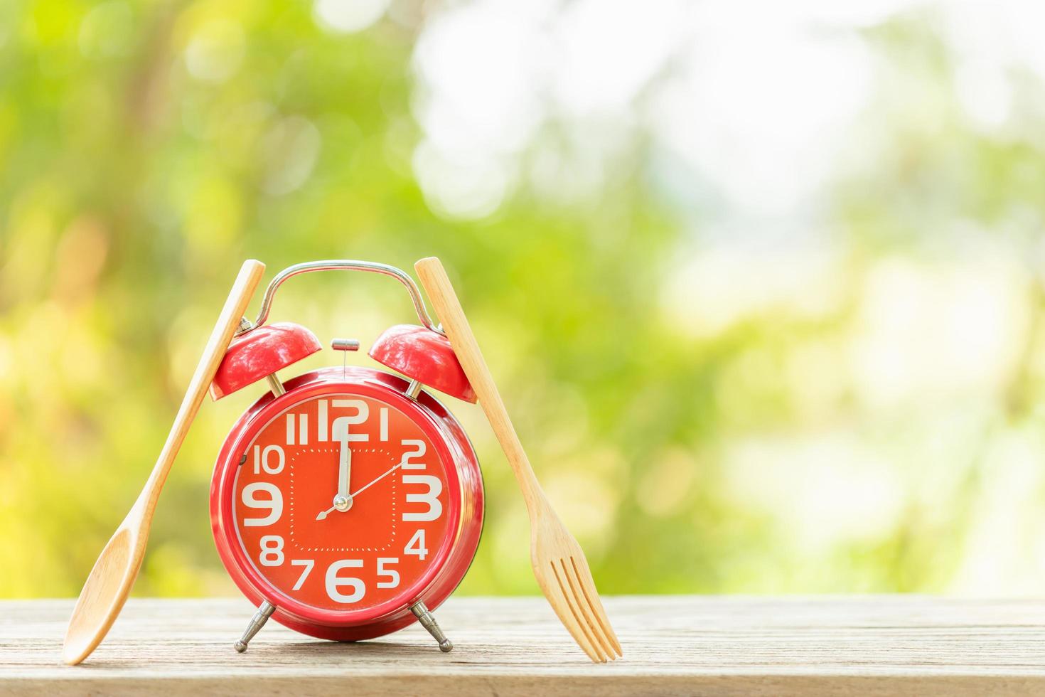 Red alarm clock, Fork, and spoon on wooden table with green outdoor nature blur background. Eight o'clock, Time for eating concept photo