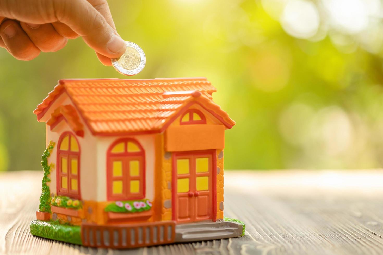 Hand put the coin to piggy bank Home Model on green nature blur background. Money savings concept photo