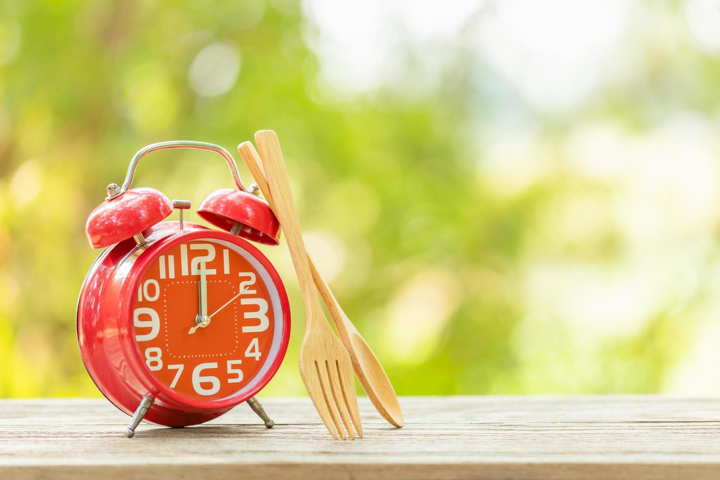 Red alarm clock, Fork, and spoon on wooden table with green outdoor nature blur background. Eight o'clock, Time for eating concept photo