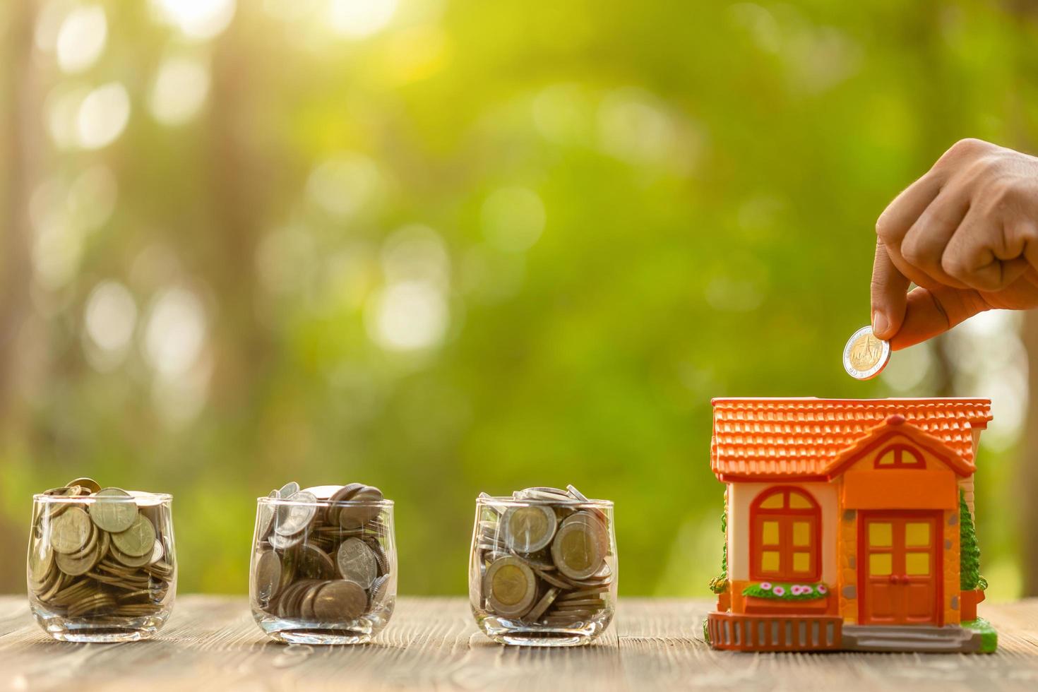 Hand put the coin to piggy bank Home Model on green nature blur background. Money savings concept photo