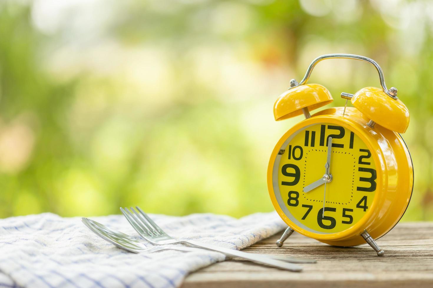 Yellow alarm clock, Fork, and spoon on wooden table with green outdoor nature blur background. Eight o'clock, Time for eating concept photo