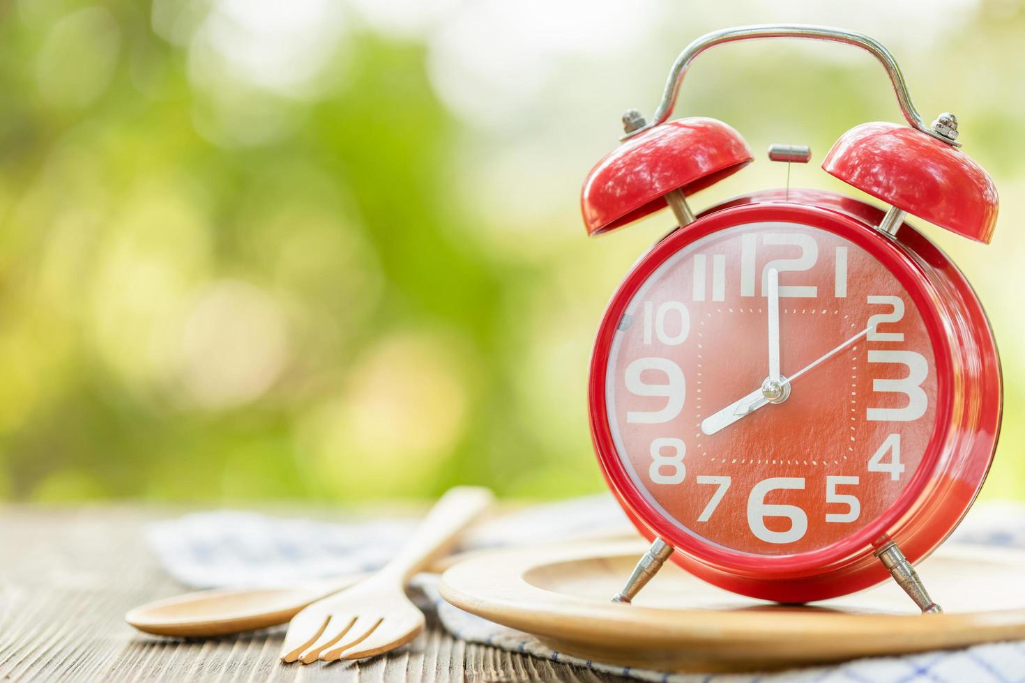Red alarm clock, Fork, and spoon on wooden table with green outdoor nature blur background. Eight o'clock, Time for eating concept photo