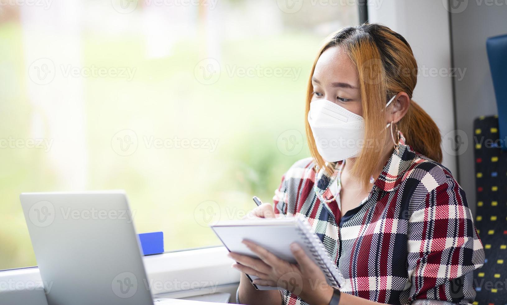 Businesswoman wearing a mask and working on laptop on a train, Asian freelance writer or designer writing emails on computer, working and smile resting on comfortable armchair near window of train. photo