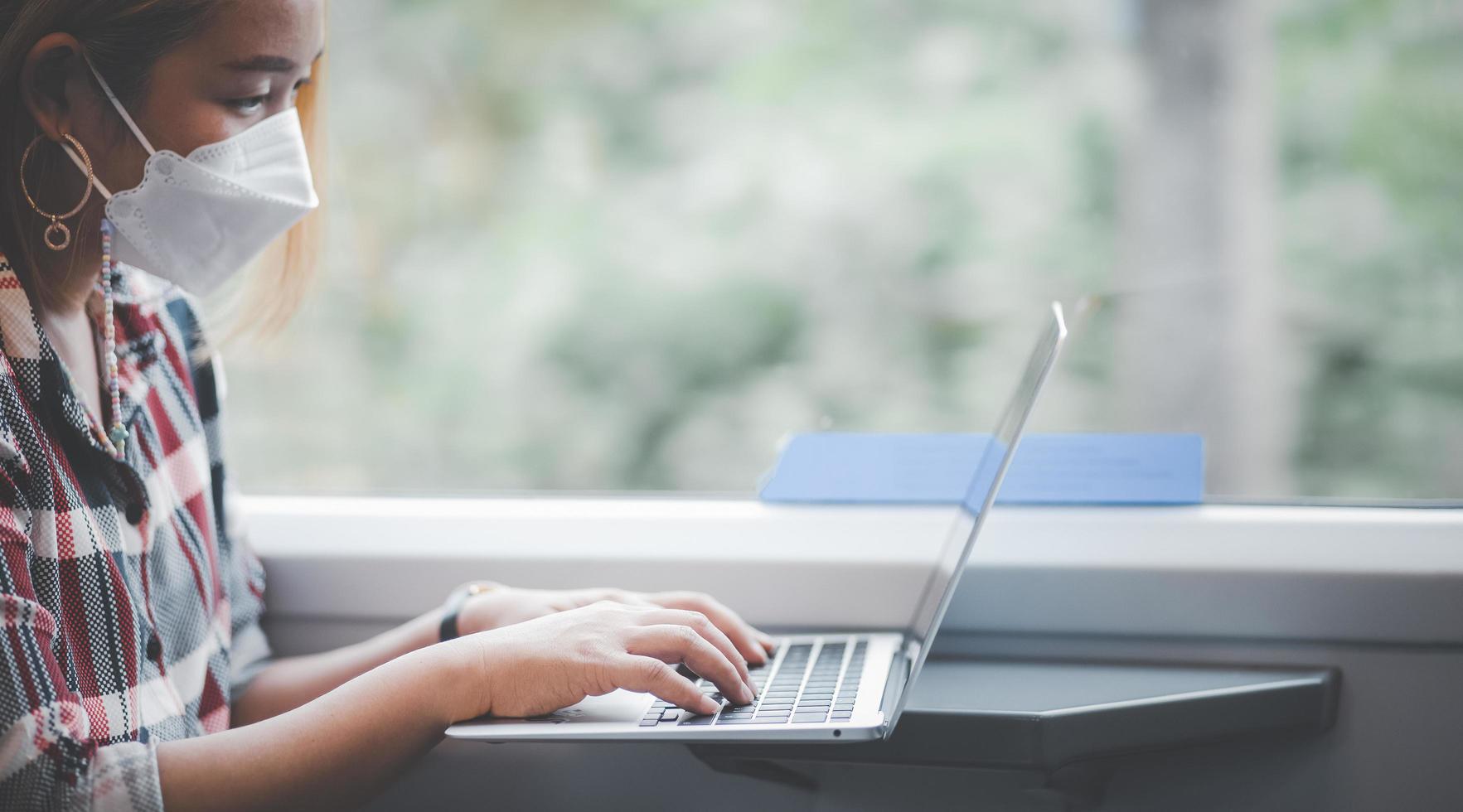 mujer de negocios con una máscara y trabajando en una computadora portátil en un tren, escritora independiente asiática o diseñadora escribiendo correos electrónicos en la computadora, trabajando y sonriendo descansando en un cómodo sillón cerca de la ventana del tren. foto