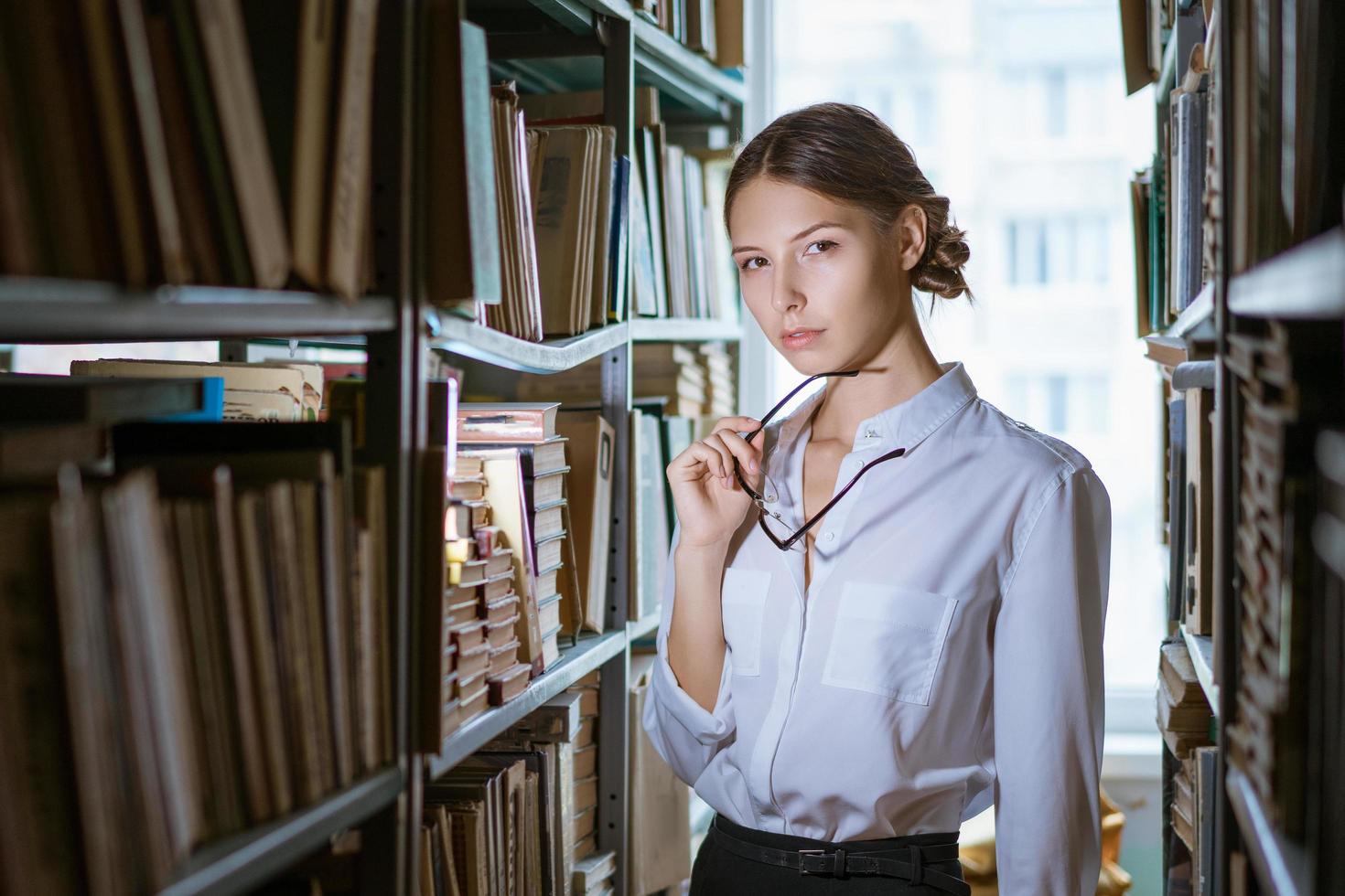 Beautiful female student  stands between the rows in the library, photo