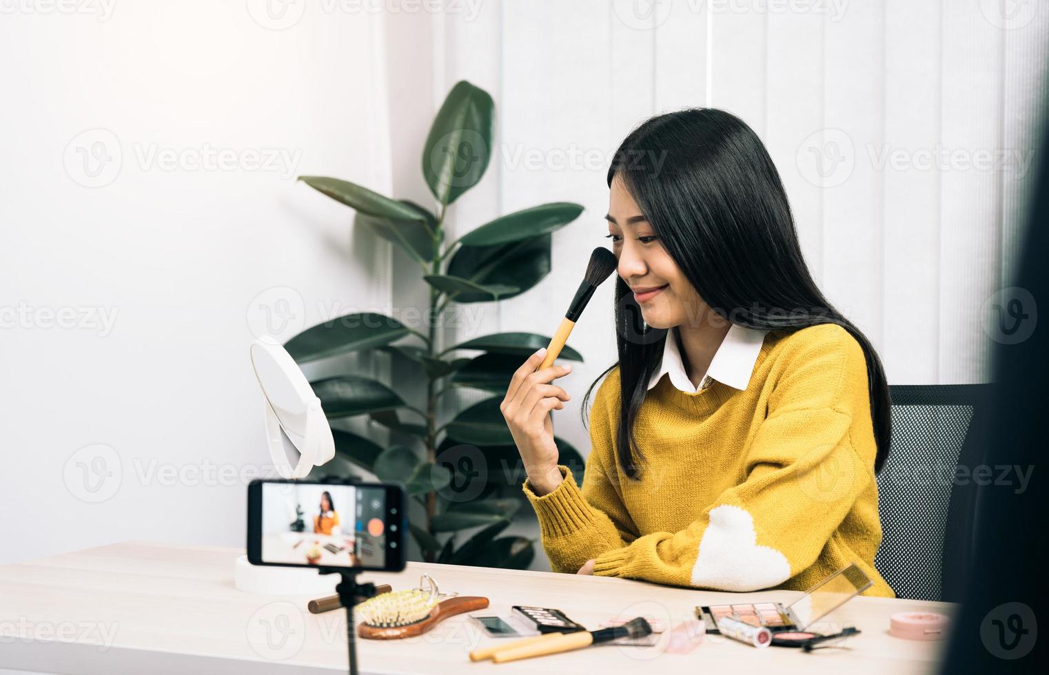 Beautiful asian woman blogger using camera phone recording vlog video live streaming and showing how to make up cosmetics at home. photo