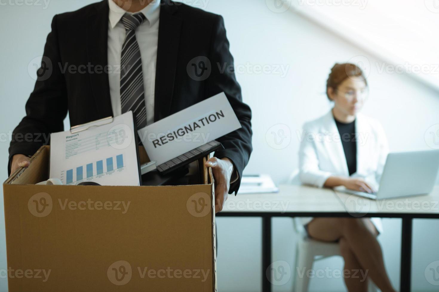 Front view with the male employee standing holding office supplies in the paper box going to submit a resignation letter while a female employee is working behind. photo