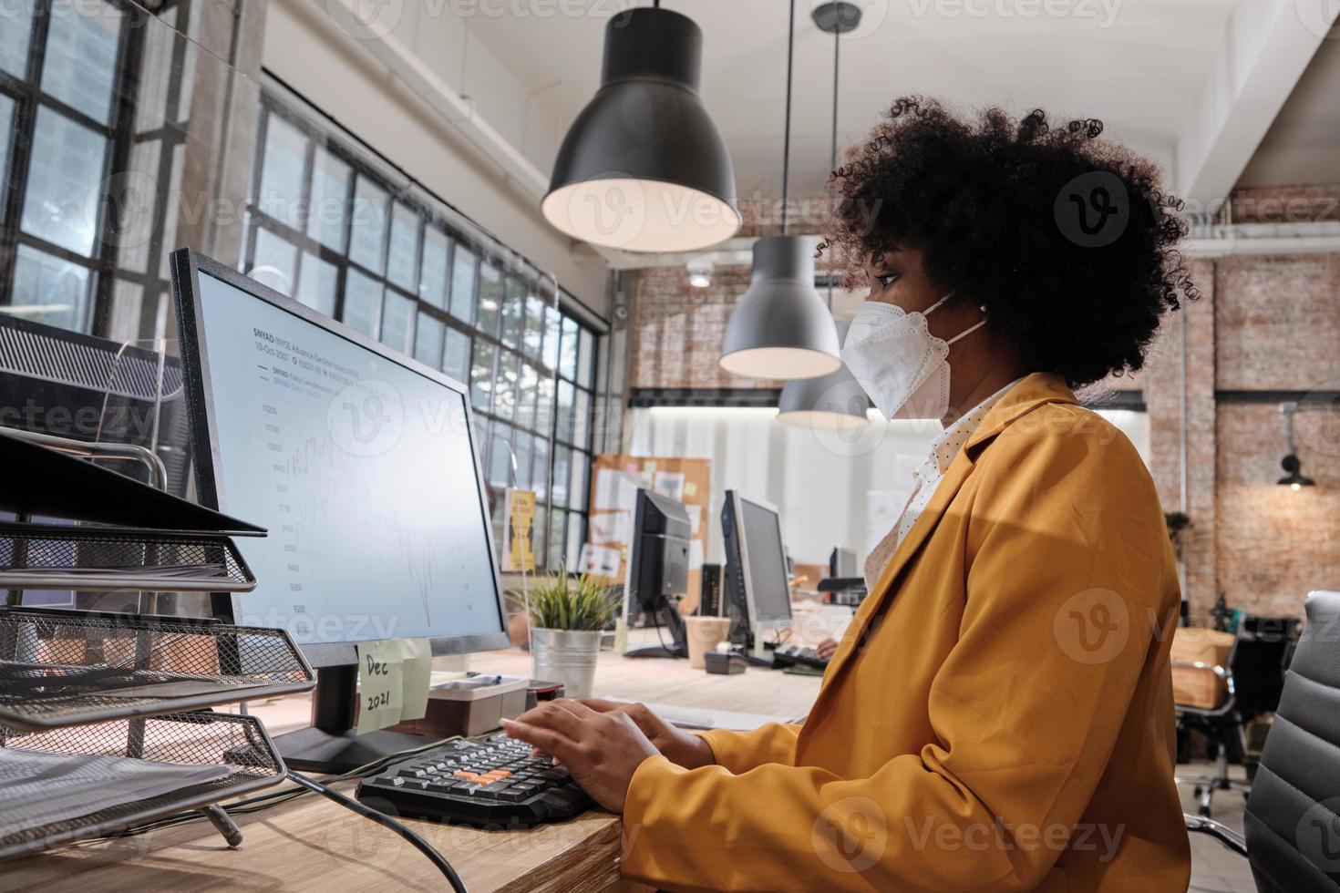 máscara facial personal femenino afroamericano que trabaja en una nueva oficina normal. protección covid-19, partición despejada, oficina de trabajo comercial, distanciamiento social para la salud pandémica, prevención de enfermedades. foto