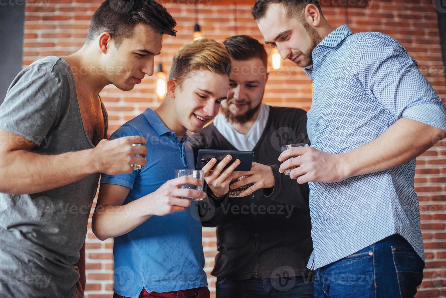 viejos amigos alegres se comunican entre sí y miran por teléfono, vasos de whisky en el pub. concepto de estilo de vida de entretenimiento. personas conectadas wifi en una reunión de mesa de bar foto