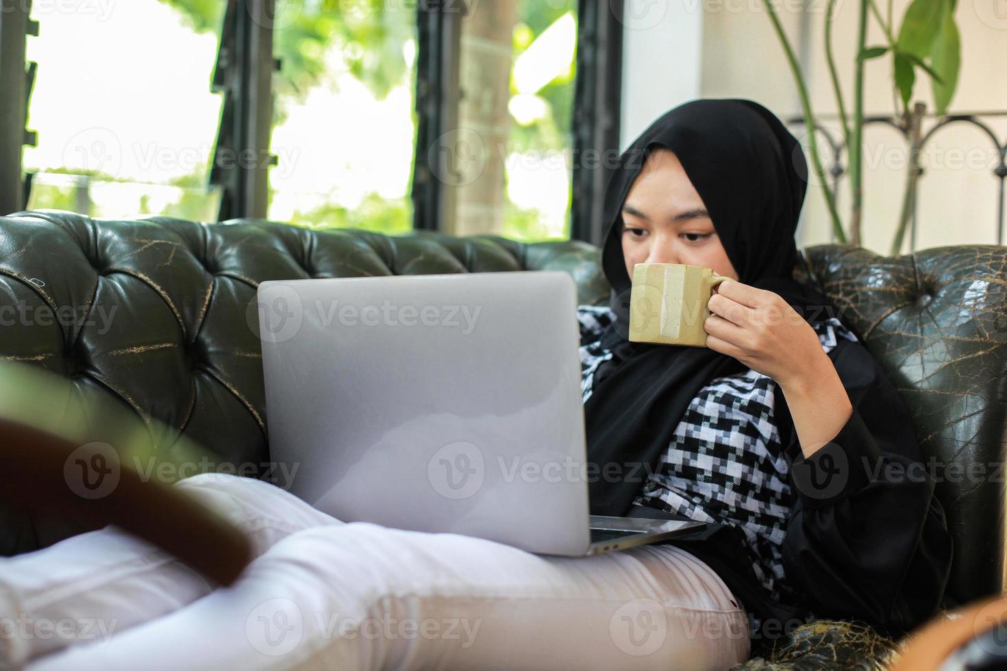 Young woman holding cup and looking at laptop while relaxing on couch photo