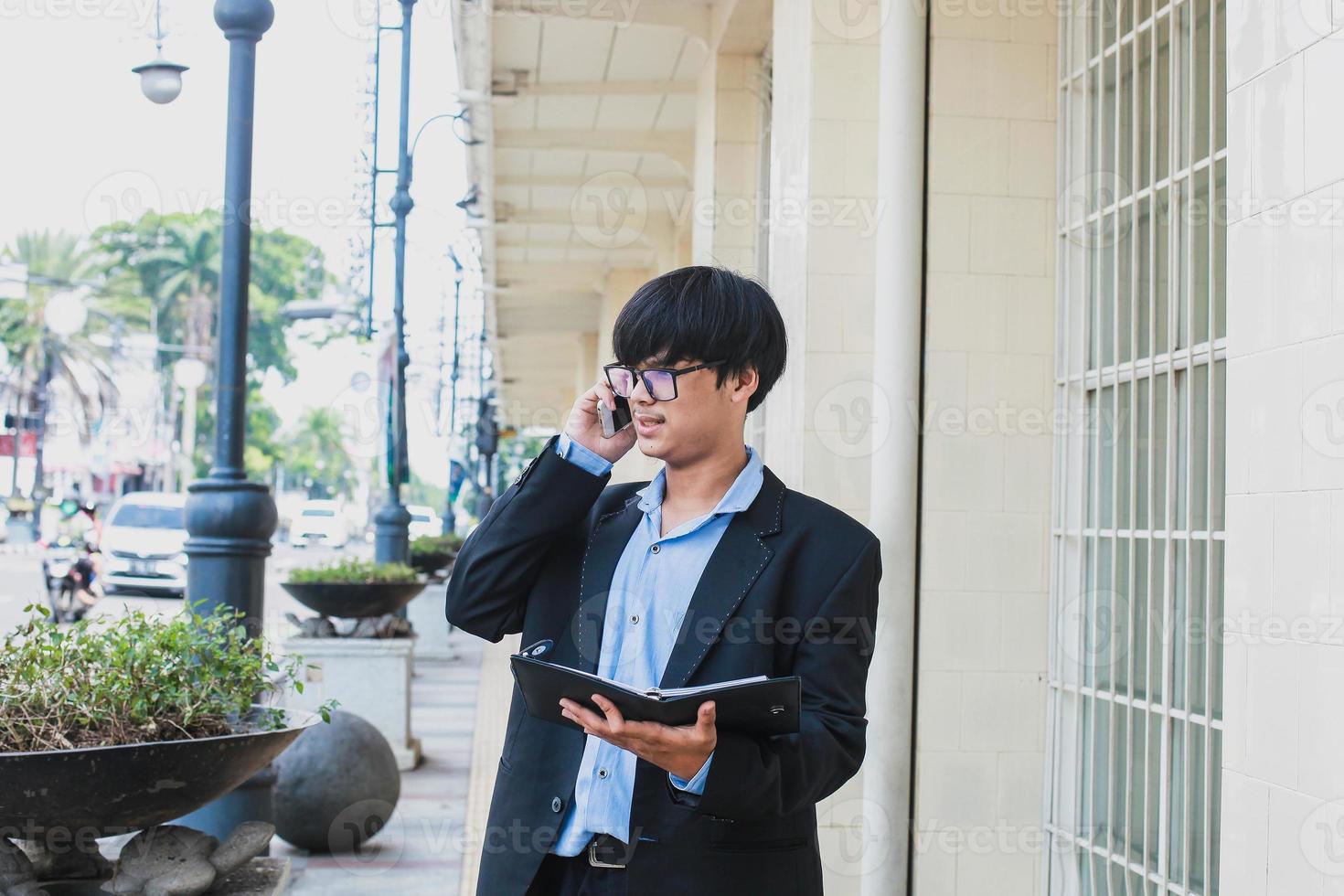 joven asiático con traje negro y anteojos llamando por teléfono mientras sostenía un cuaderno y miraba en la calle foto