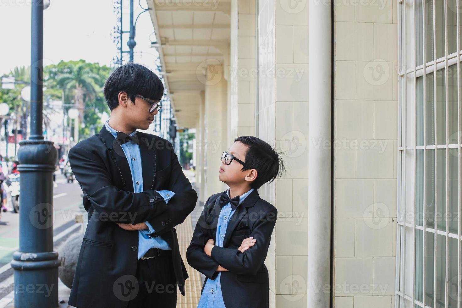 Two teenager wearing glasses, black ribbon and vintage black suit posing in front of the camera while crossed arm and looking at each other photo