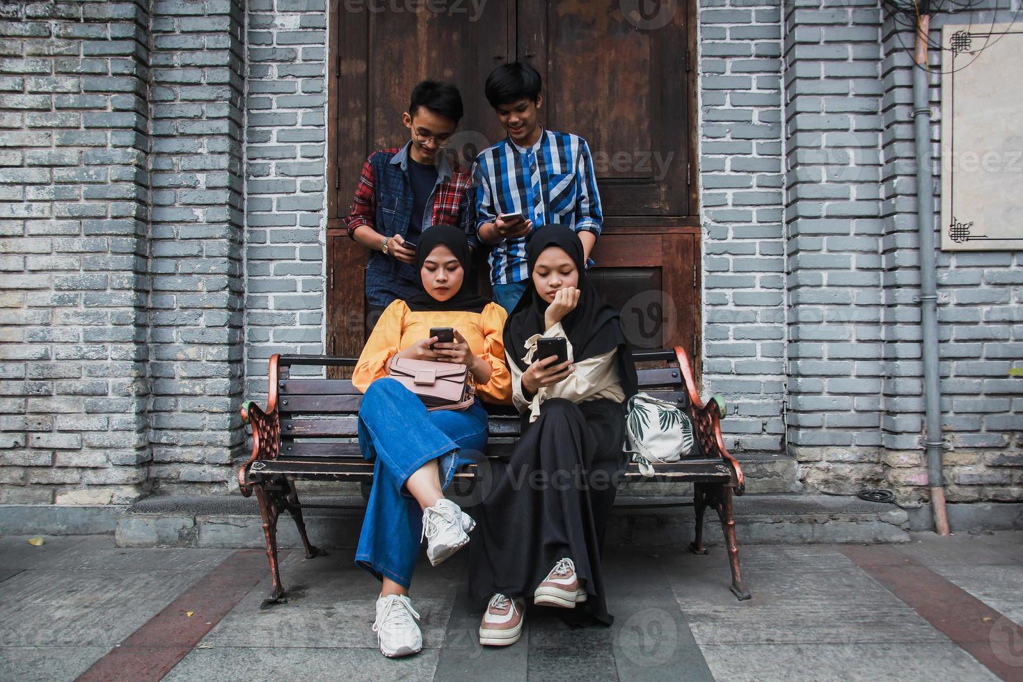 Four young friends in outdoors and looking at mobile phone. Two female sitting on a bench looking mobile phone with a frowny face and two male standing behind them smiling and holding smartphone photo