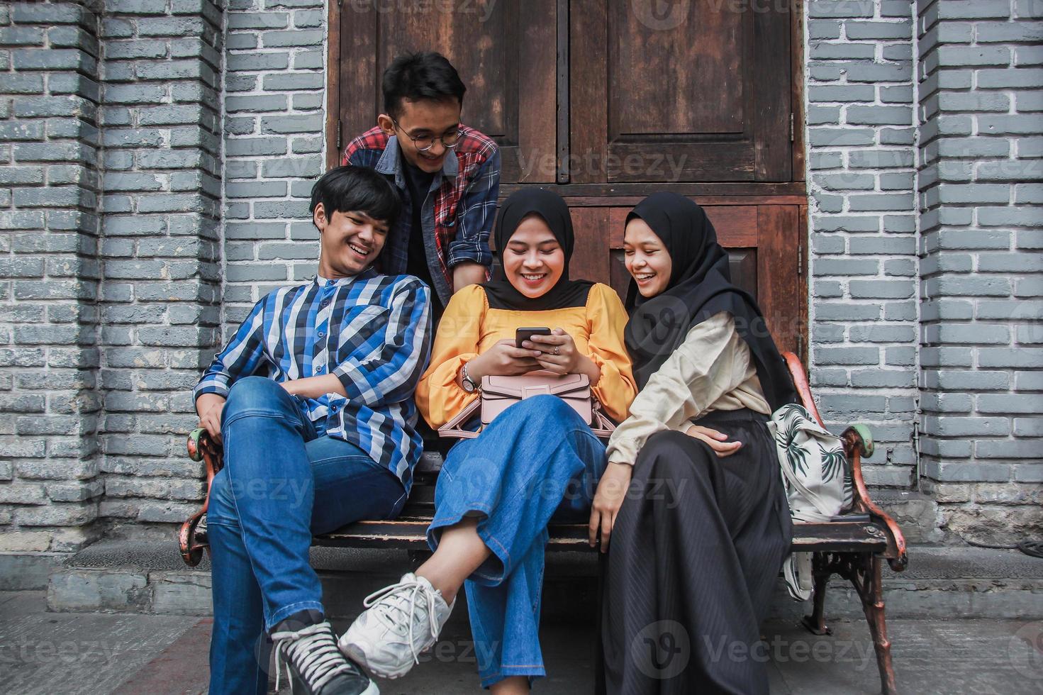 Four young friends sitting outdoors and looking at mobile phone. Group of people sitting on a bench and watching video smiling and having fun together photo