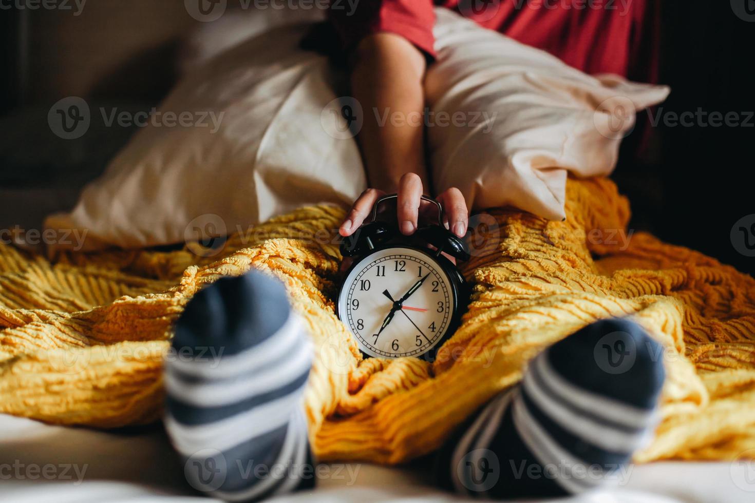 gente durmiendo con reloj en la mano mostrando las 7 en punto con el pie en el calcetín debajo de la manta foto