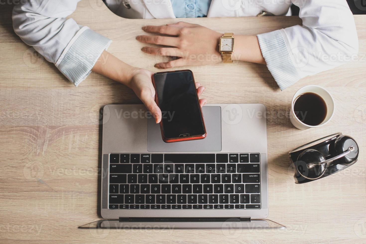 Top view of woman hand are holding and using blank screen smartphone with laptop, coffee and clock on a wooden office desk photo