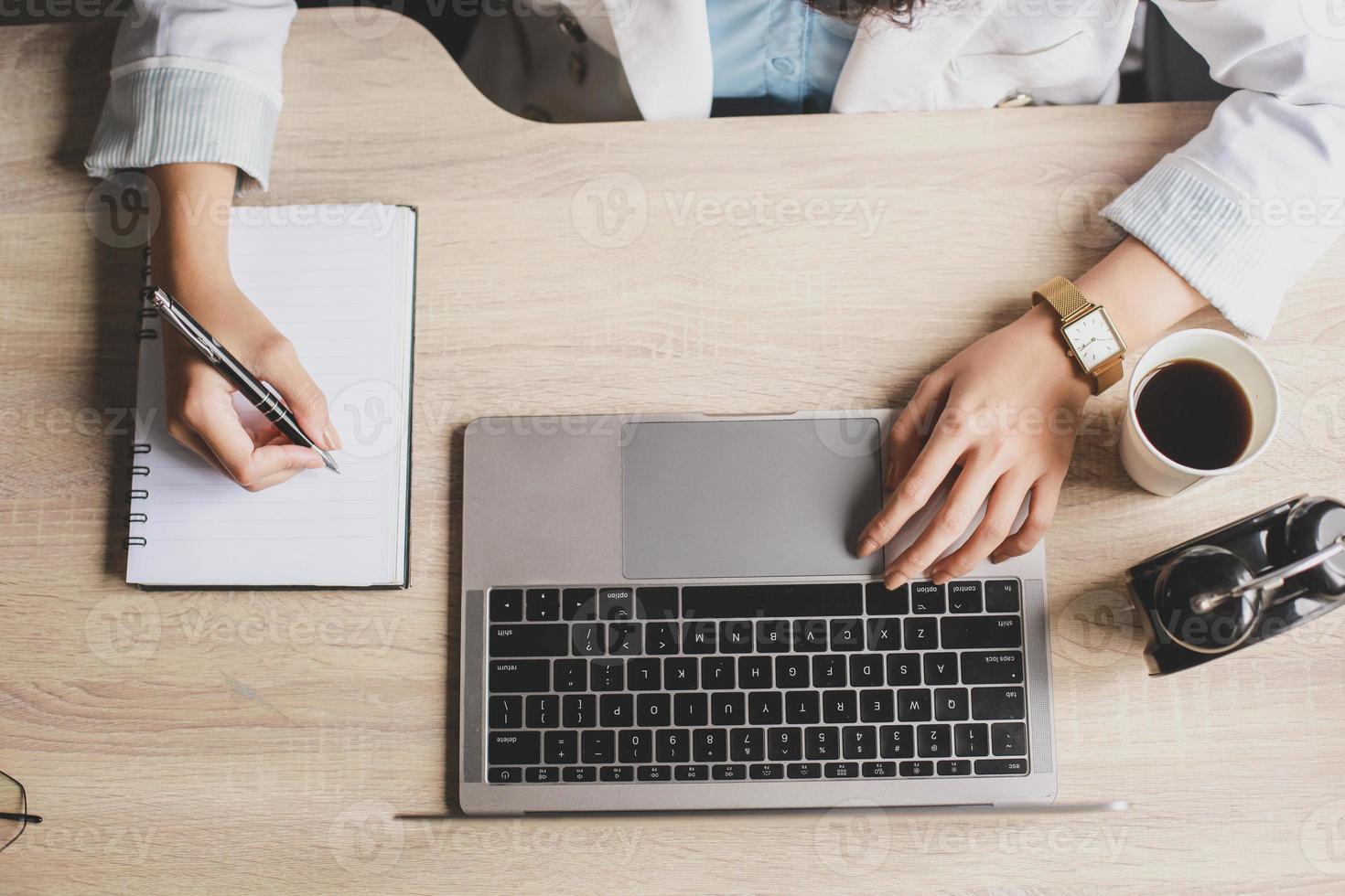 Top view of woman hand are writing on the book and using a laptop computer with coffee and clock on a wooden office desk photo