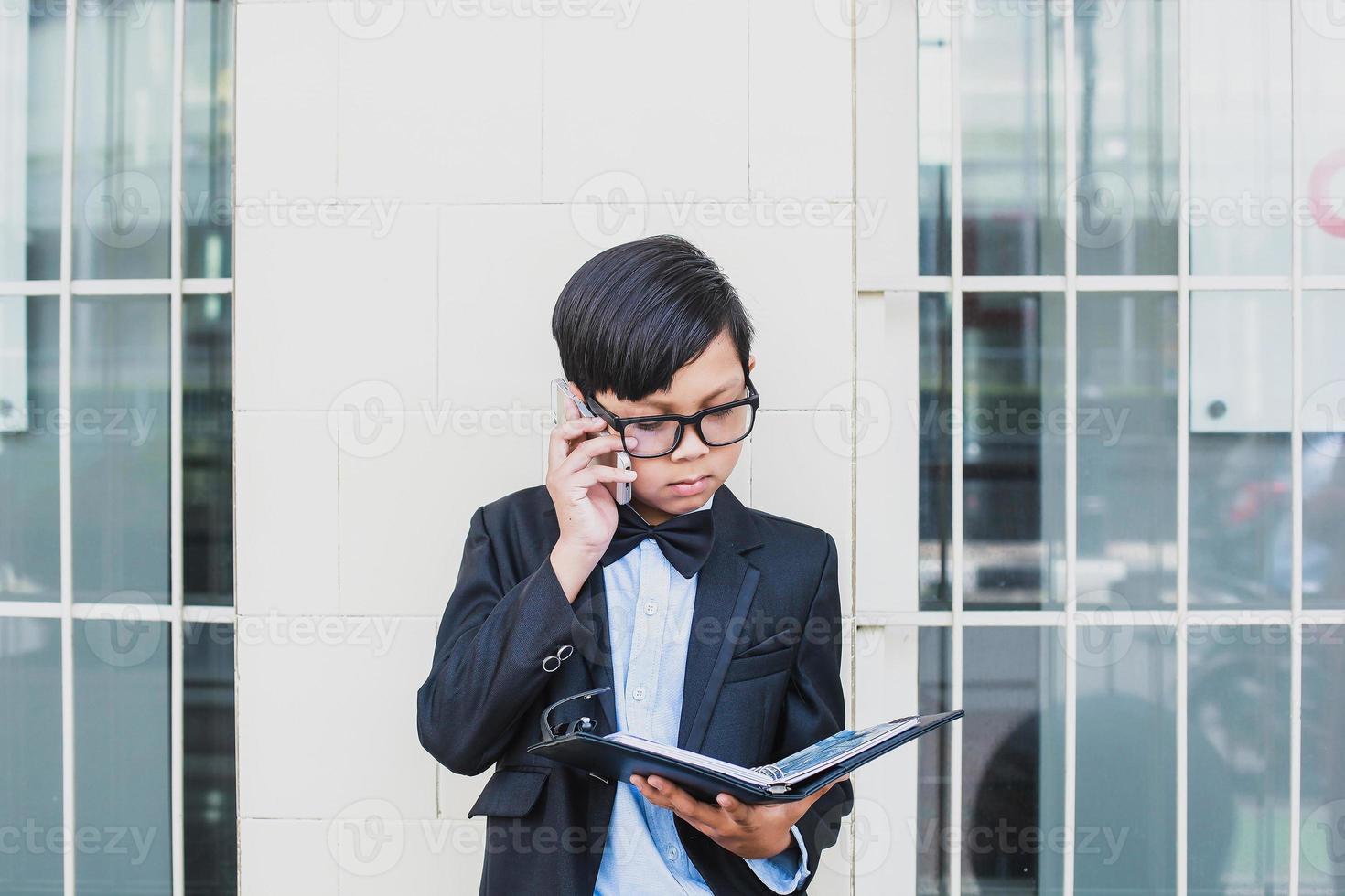 niño asiático con traje negro vintage y anteojos haciendo una llamada por teléfono mientras mira un cuaderno foto