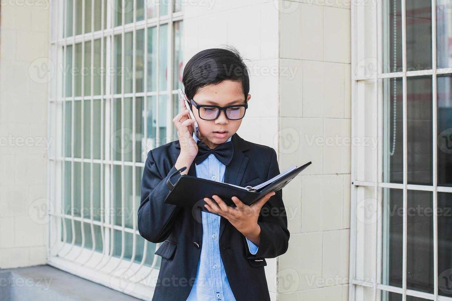 niño asiático con traje negro vintage y anteojos haciendo una llamada por teléfono mientras lee un libro de notas foto