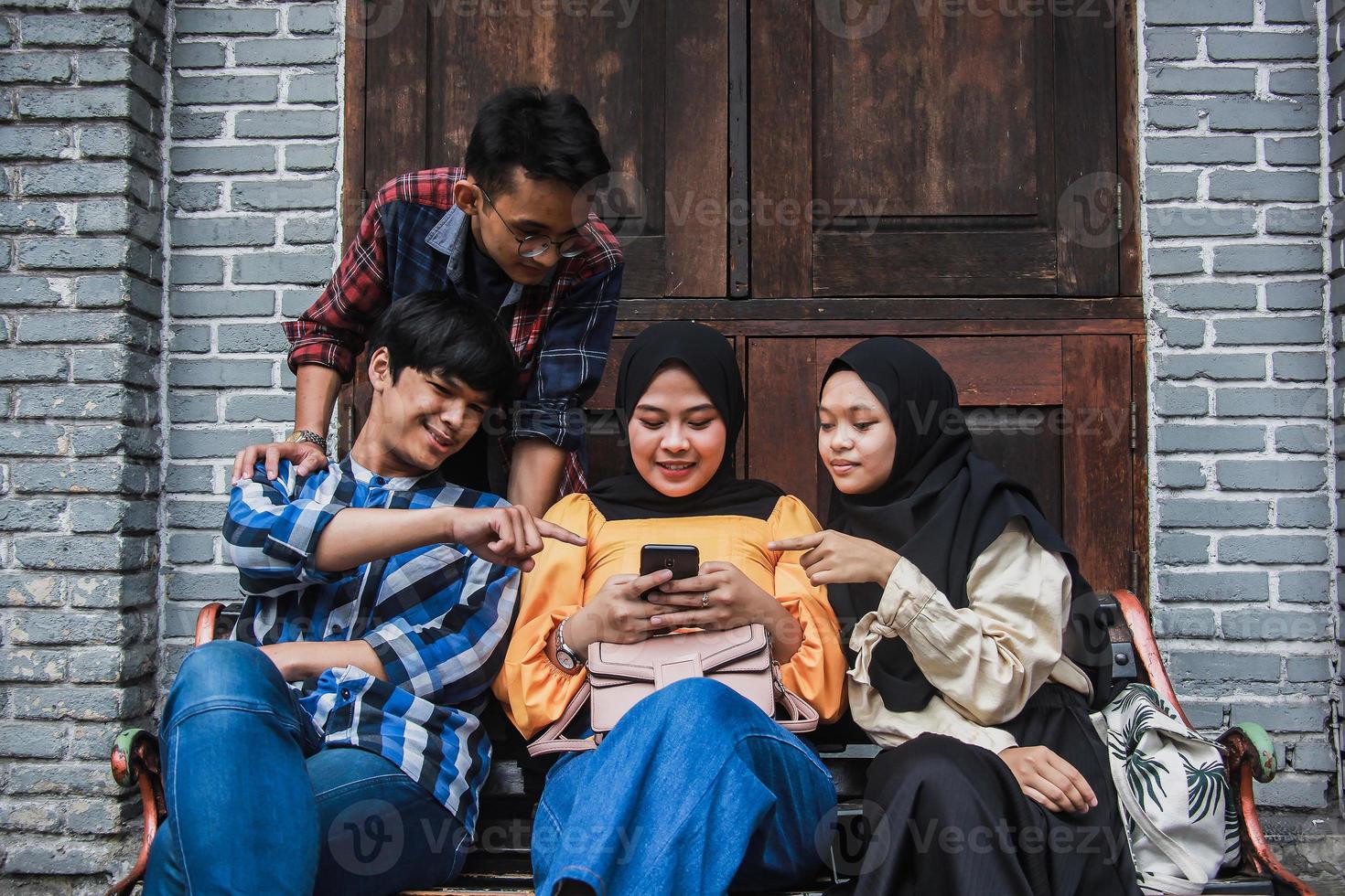 Four young friends sitting outdoors and looking at mobile phone. Group of people sitting on a bench and watching video smiling and having fun together photo