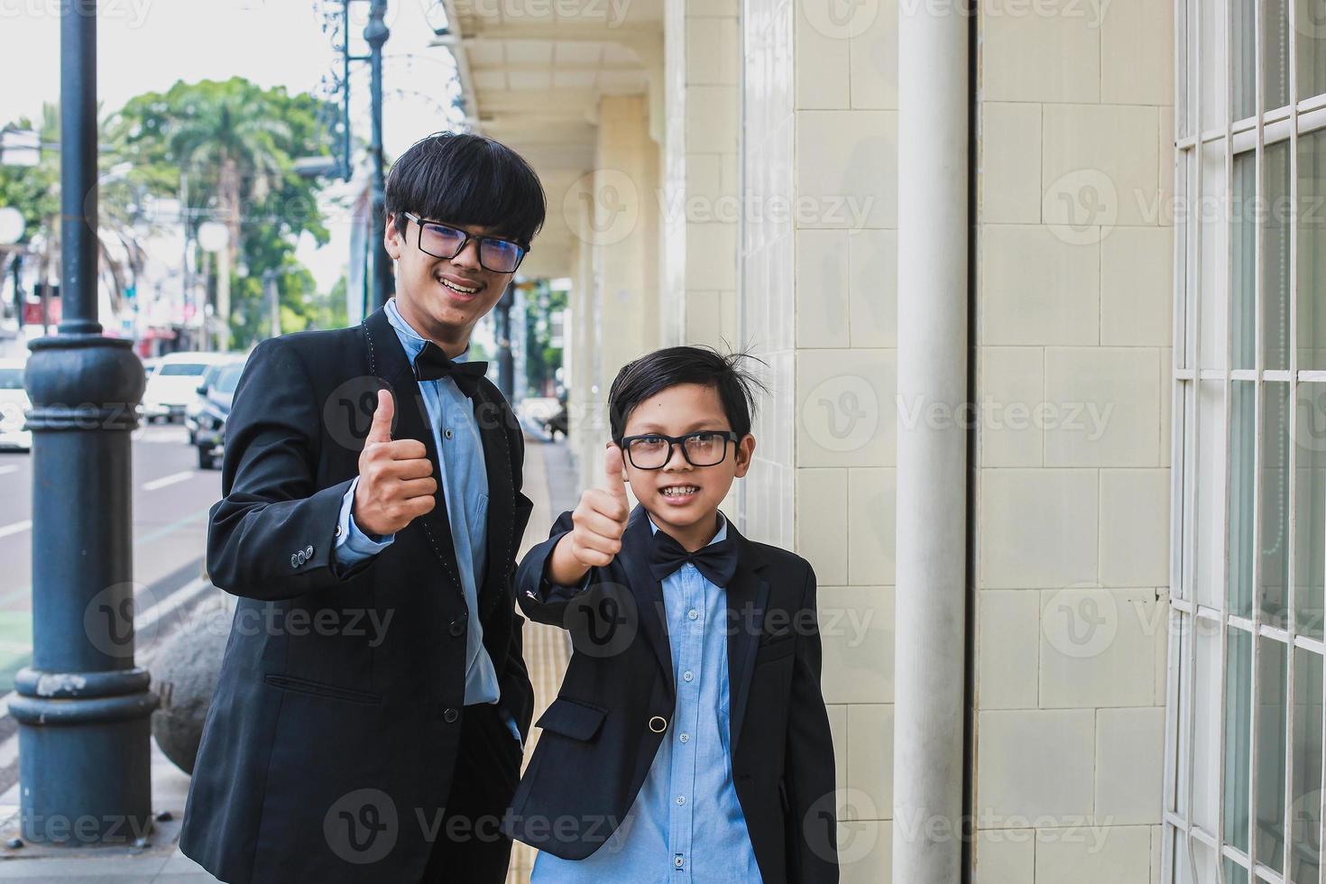 Two teenager wearing vintage black suit posing and smiling into the camera while showing thumb up or like gesture photo