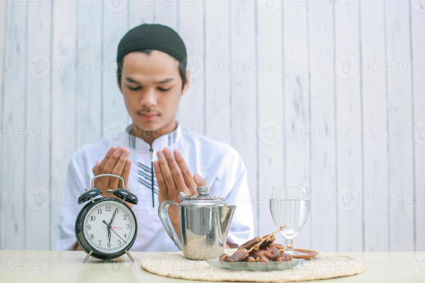 Blurry photo of young muslim man praying to God at iftar time, selective focus on clock. Traditional Ramadan, iftar meal. Ramadan kareem fasting month concept