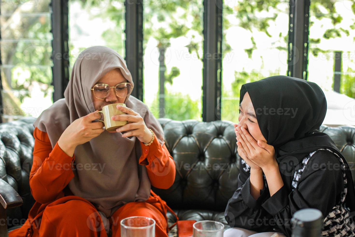 Two muslim women sitting on couch, drinking coffee and joking photo