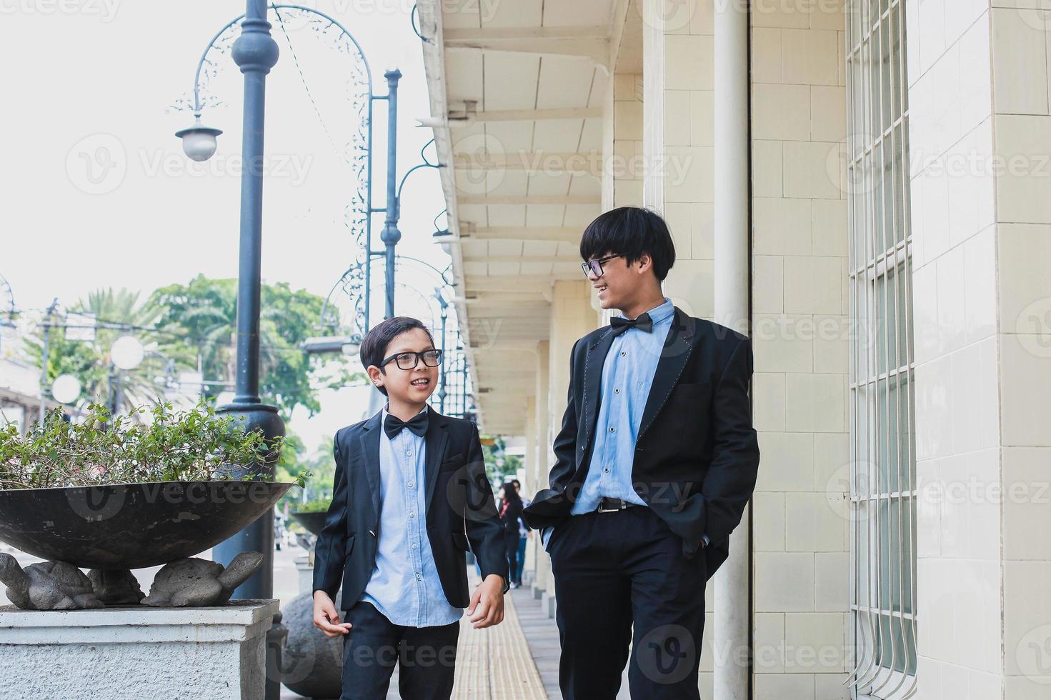 Two teenager wearing vintage black suit walking together while joking at sidewalk photo