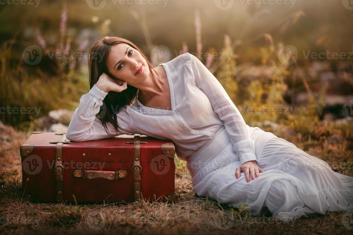 young woman sitting in the field with suitcase photo