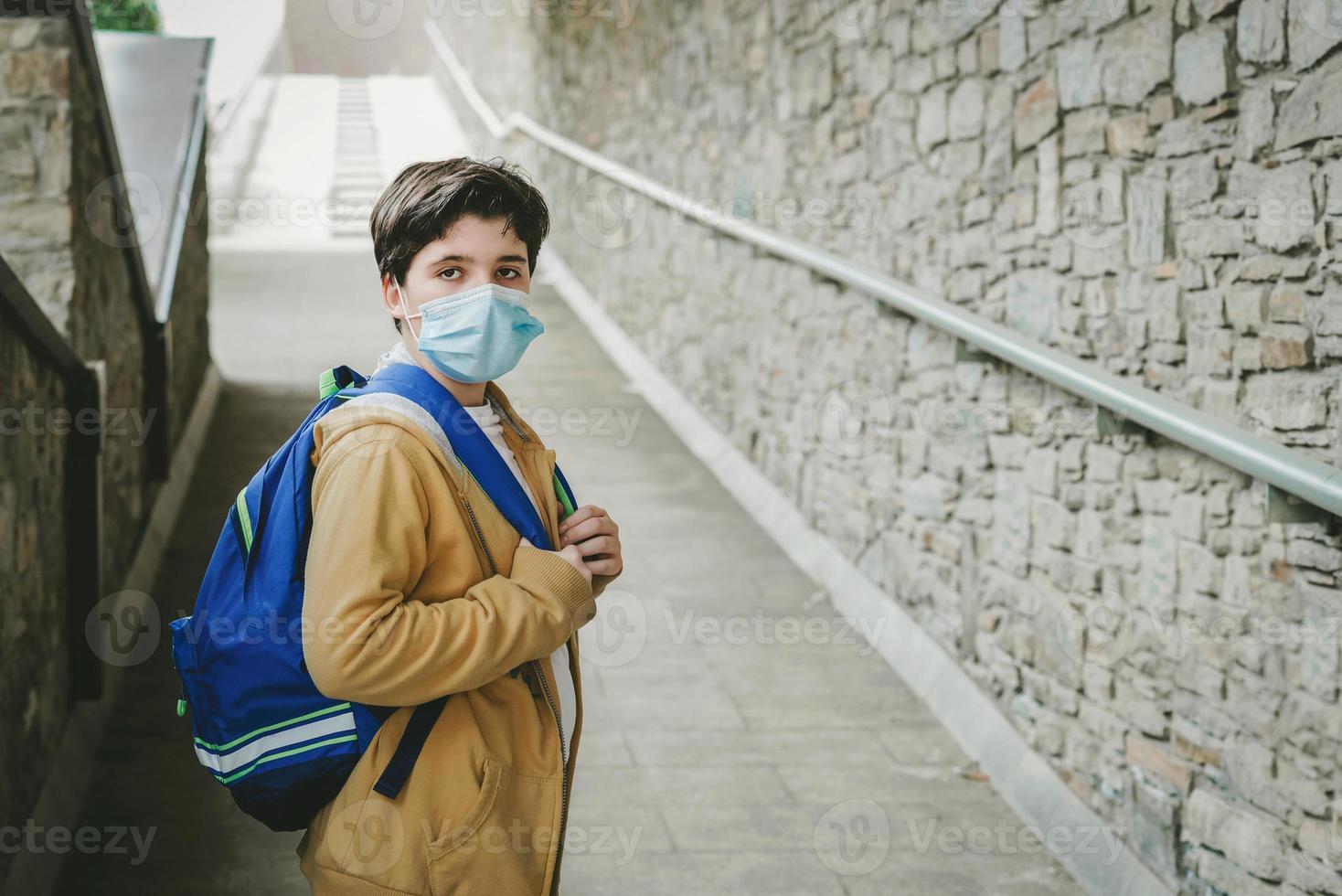 retrato de un niño con máscara médica y mochila esperando en la puerta de la escuela foto