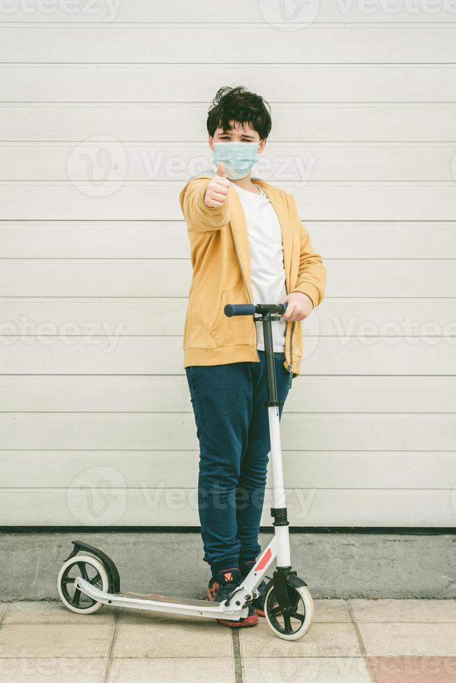 niño feliz usando máscara médica con su scooter en la ciudad foto